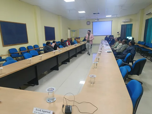 Presenter walking along a long seminar-hall table while participants listen